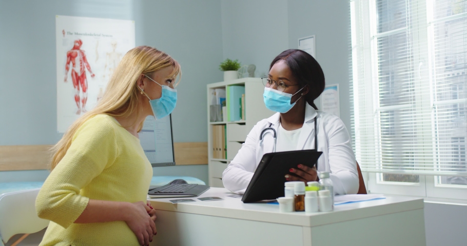 Medium shot of pregnant woman visit african american gynecologist in medical mask at hospital. Doctor showing tablet to female and consulting. Pregnancy and maternity healthcare concept. | Shutterstock HD Video #1066286521