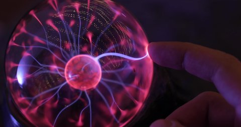 Hand touching with finger electric plasma ball with luminous flames in the dark room. Tesla glass sphere in action with smooth magenta-blue rays.
