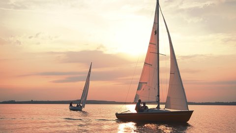 A cinematic scene of two small sailing yacht boats sailing along the golden calm waters on lake, river, sea or ocean. Beautiful sunset or sunrise, relaxation, calmness, sailing, sailors. Luxury living