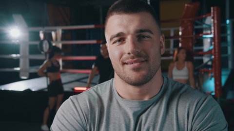 Sport and trainings concept. Portrait of tired muscular man boxer looking at camera with pleasure smile at the dark room with boxing ring at the background. Sportsman relaxing after physical training