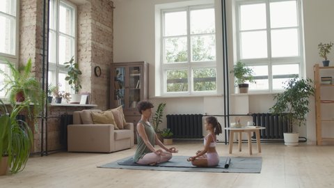 Wide shot of young woman and little girl in sportswear sitting together in lotus pose on mat and meditating in living room at home