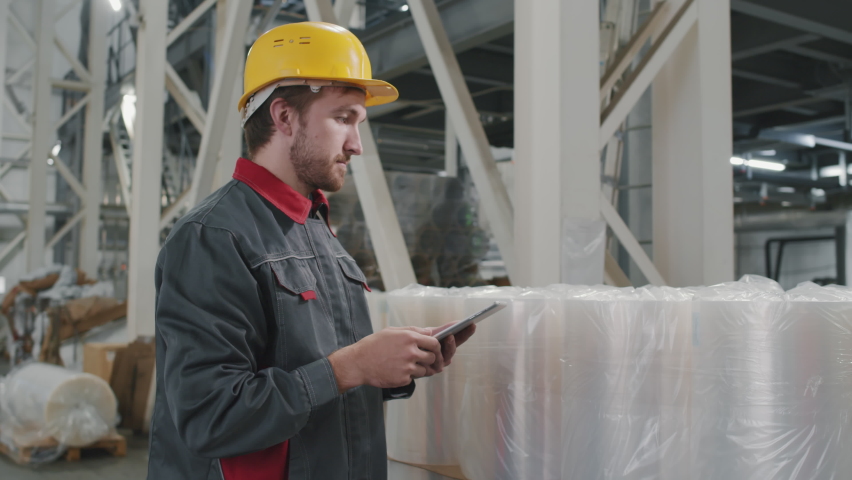 Side-view slow-motion medium shot of caucasian workman wearing safety helmet checking produced rolled polyethylene making notes on digital tablet | Shutterstock HD Video #1066311427
