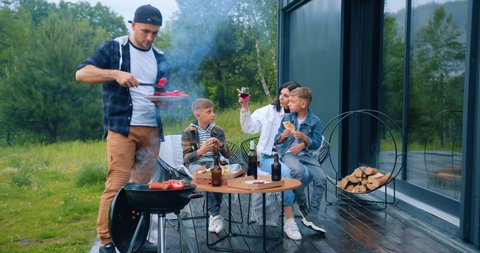 Family outdoors picnic concept where attractive positive father cooking vegetables and sausages on grill when children talking with mother around the table