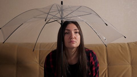 Upset brunette lady hides from water flowing from upstairs neighbors under transparent umbrella sitting on sofa in dark room close-up. Concept of flooding the apartment and property insurance