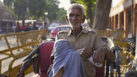 Frontal shot of extremely old Indian man with white hair sitting comfortably with crossed legs on a cycle rickshaw parked by a roadside in a busy market place looking at the camera