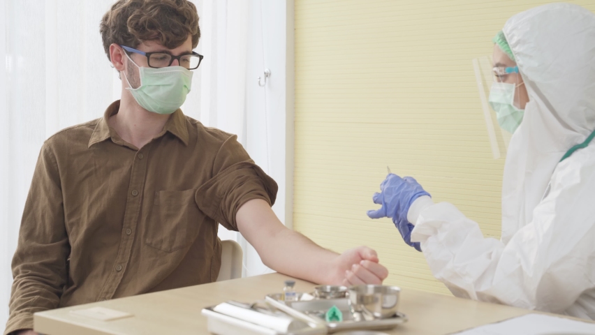 A white young female doctor sitting in a bright hospital examination room  wearing PPE,mask,protective device is taking out a newly develop covid19 vaccine to inject to a young white male patient | Shutterstock HD Video #1066319359