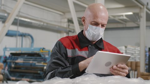 Medium slowmo close-up of caucasian factory workman in protective mask checking quality of produced polyethylene making notes on digital tablet