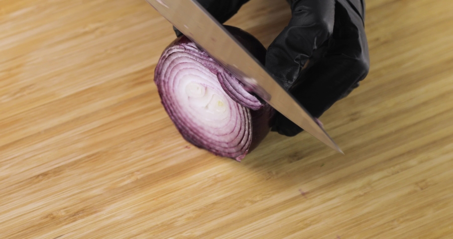 Cook in gloves is preparing vegeterian healthy food, salad. Chef is cooking cutting fresh red onion on wooden board, hands closeup. Slicing red onion in kitchen using knife.