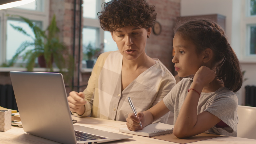 Tilt up shot of young mother with short curly hair sitting at desk beside her frustrated little daughter and explaining something to her while helping her with homework | Shutterstock HD Video #1066325323