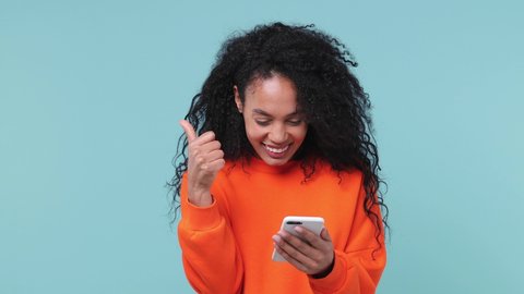 Shocked amazed fun young african woman in orange sweatshirt hold using mobile cell phone typing browsing say wow yes just found out great big win news doing winner gesture isolated on blue background