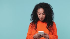 Young african woman with in orange sweatshirt get video call using mobile cell phone doing selfie videoconference talking conducting pleasant conversation greets with hand isolated on blue background