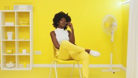 Portrait of black woman, young and beautiful model, sitting on yellow chair next to a fan, looking at camera confidently, trying to chill out, posing on camera, Slow motion.