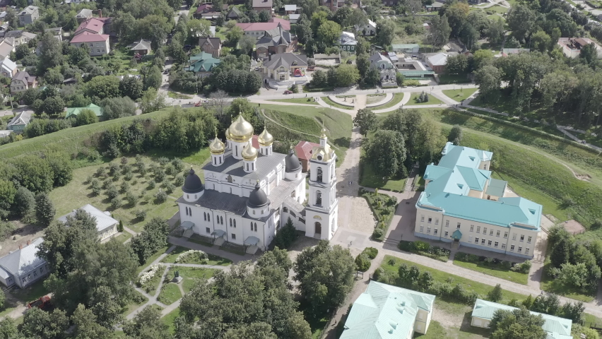 Dmitrov, Russia. The Cathedral of the Assumption of the Blessed Virgin Mary stands within the Dmitrov Kremlin, an early 16th-century architectural monument commissioned by Prince Yuri Ivanovich and constructed over 22 years. The white stone cathedral features a central bell tower and multiple gilded and dark domes crowned with Orthodox crosses, surrounded by historic buildings with turquoise roofs and remnants of earthen fortifications. The aerial perspective reveals the cathedral