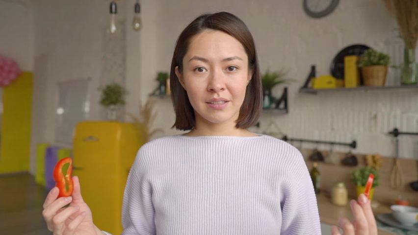 Funny mix race asian girl dancing and enjoying cooking meal at home, having fun while preparing salad make funny faces cover eyes with red paprika circles. Healthy cooking vegan meal cocept