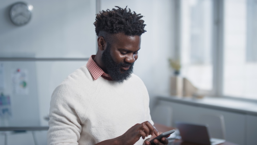 Slow motion portrait shot footage of young adult African American guy standing in office room using his smartphone then looking away | Shutterstock HD Video #1066343161