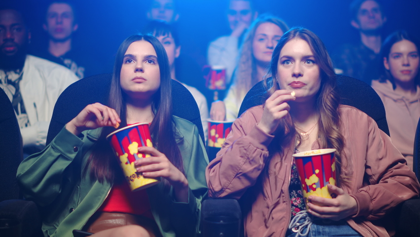 Young women watching film in dark hall. Surprised girl pointing on screen in cinema. Attractive female friends speaking in movie theater. Young girlfriends eating popcorn. | Shutterstock HD Video #1066344493