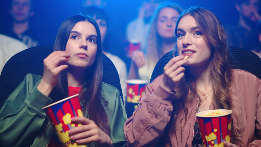 Young women watching film in dark hall. Surprised girl pointing on screen in cinema. Attractive female friends speaking in movie theater. Young girlfriends eating popcorn. Royalty-Free Stock Footage #1066344493