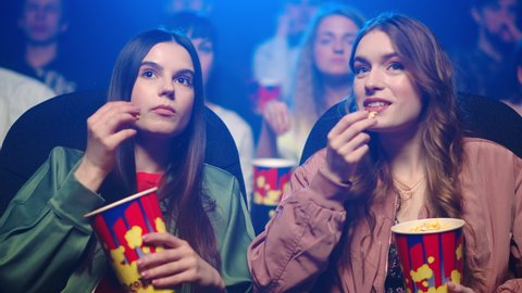 Young women watching film in dark hall. Surprised girl pointing on screen in cinema. Attractive female friends speaking in movie theater. Young girlfriends eating popcorn.