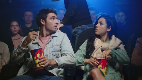 Young couple getting irritated with man behind in movie theater. Angry man looking back in cinema. Beautiful couple eating popcorn indoor. Group of people watching movie in dark hall.