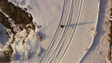 Woman on snowy road, aerial top view.  Field covered by snow. 
Alone girl in dark coat walking on snowy trail, aerial top down footage. tire track on the snow. 