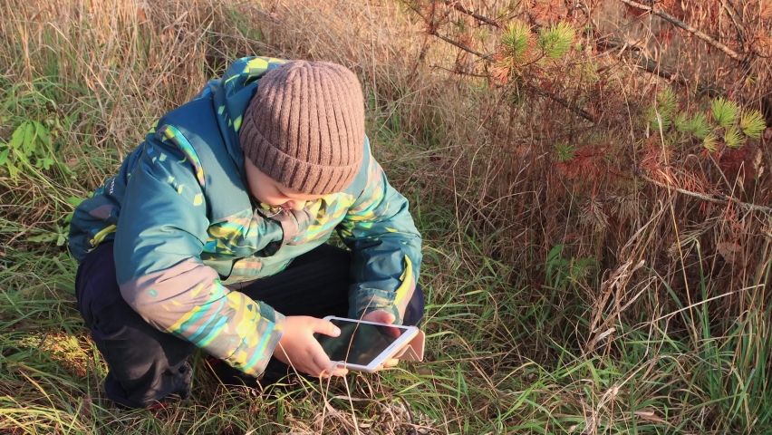 A boy squatting uses the Internet online on a tablet and looks. Walk in the park in autumn. Camping on a hike. Healthy lifestyle concept. At sunset, in the evening, warm soft light.