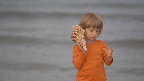 Portrait of a blond hair child with blue eyes walks on the beach holding a shell to his ear to hear the sea music.