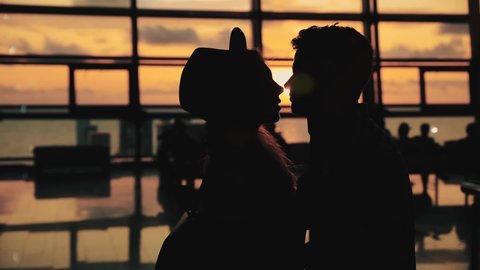 Silhouette of man and a woman kissing in airport terminal after arrival. Happy couple meet after long separation. Sunset panoramic window at background.