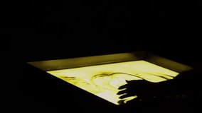 Girl caucasian hand sprinkles sand on a yellow glowing table, sand painting