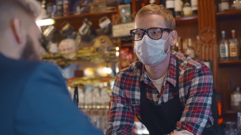 Back view of man client talking to bartender wearing safety mask and gloves. Drunk lonely businessman sitting at bar counter and talking to barman in protective mask.