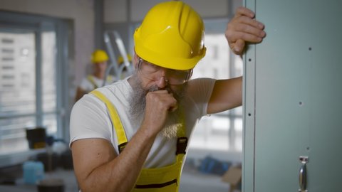 Senior construction worker sick and coughing in renovating apartment. Portrait of aged builder male in yellow overall and hardhat feeling unwell and leaning on wall at construction site.