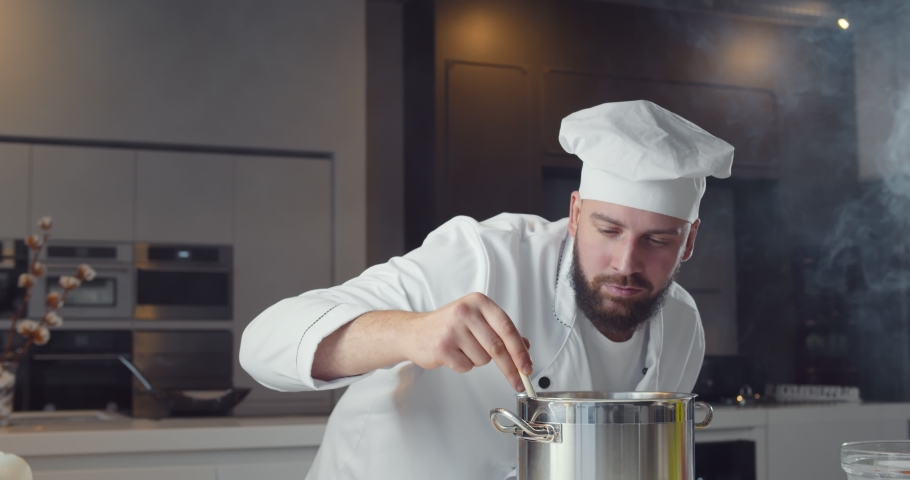 Professional chef stirring soup with spoon in restaurant kitchen. Portrait of handsome male cook in white coat and hat uniform cooking sauce in pan. Royalty-Free Stock Footage #1066352248