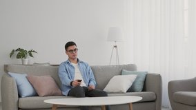 Young guy in eyeglasses watching tv at home, sitting on sofa and switching channels, slow motion