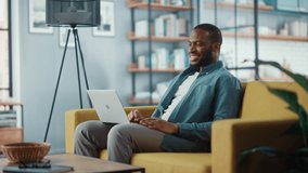 Handsome Black African American Man Having a Video Call on Laptop Computer while Sitting on a Sofa in Living Room. Freelancer Working From Home and Talking to Colleagues and Clients Over the Internet.