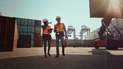 Multiethnic Female Industrial Engineer with Tablet Computer and Male Foreman Worker in Hard Hats and Safety Vests Walk in Container Terminal. VFX Double Girder Gantry Cranes Work in the Background.