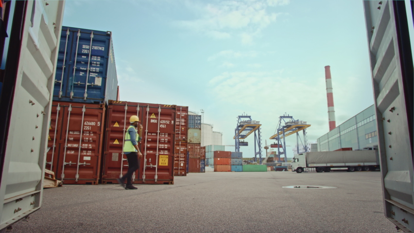 Forklift Driver Loading a Shipping Container with a Pallet with Boxes in Logistics Terminal. Female Industrial Supervisor Helping the Process. VFX Double Girder Gantry Cranes Work in the Background. Royalty-Free Stock Footage #1066358572