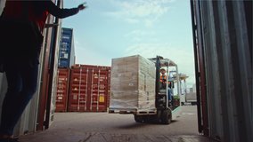 Forklift Driver Loading a Shipping Container with a Pallet with Boxes in Logistics Terminal. Female Industrial Supervisor Helping the Process. VFX Double Girder Gantry Cranes Work in the Background. - Powered by Shutterstock - Get 15% off with code: PIKWIZARD15