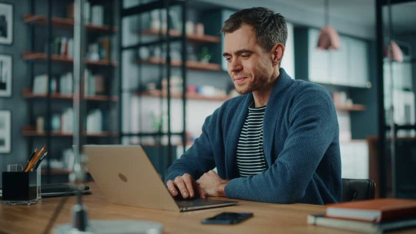 Handsome Caucasian Man Working on Laptop Computer while Sitting Behind Desk in Cozy Living Room. Freelancer Working From Home. Browsing Internet, Using Social Networks, Having Fun in Flat.