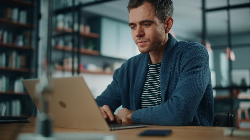 Handsome Caucasian Man Working on Laptop Computer while Sitting Behind Desk in Cozy Living Room. Freelancer Working From Home. Browsing Internet, Using Social Networks, Having Fun in Flat.