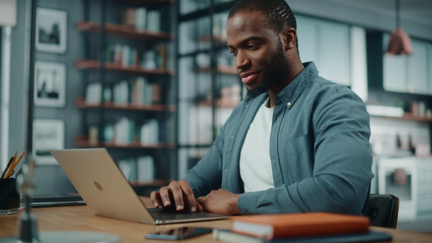 Handsome Black African American Man Working on Laptop Computer while Sitting Behind Desk in Cozy Living Room. Freelancer Working From Home. Browsing Internet, Using Social Network, Having Fun in Flat.