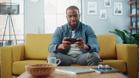Happy Black African American Man Having a Playing Video Game on Smartphone App while Sitting on a Sofa in Living Room. Excited Person of Color Resting at Home and Having Fun Over the Internet.