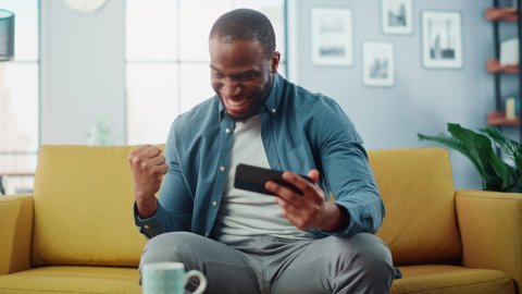 Happy Black African American Man Having a Playing Video Game on Smartphone App while Sitting on a Sofa in Living Room. Excited Person of Color Resting at Home and Having Fun Over the Internet.