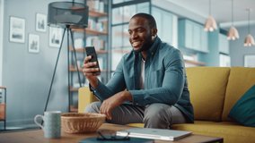 Excited Black African American Man Having a Video Call on Smartphone while Sitting on a Sofa in Living Room. Happy Man Smiling at Home and Talking to Colleagues and Clients Over the Internet.