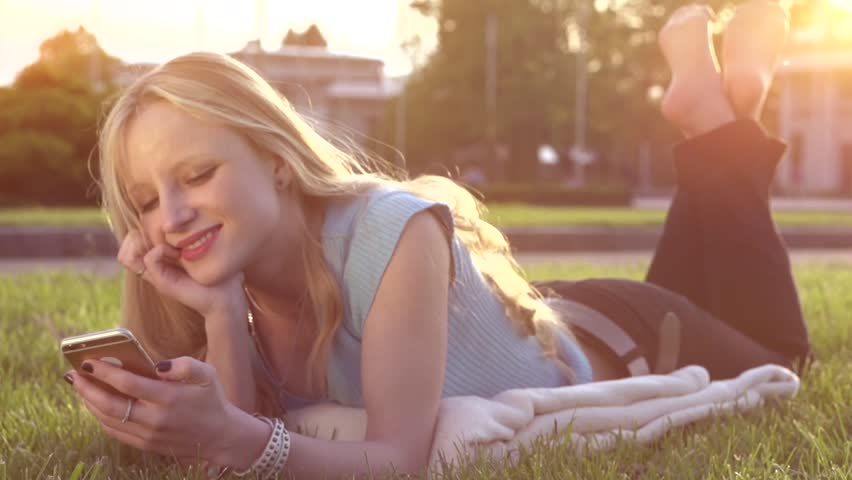 A beautiful young woman talking by mobile phone. Girl is lying on the green grass in the city park. Slow motion 240 fps. High speed camera shot. Full HD 1080p. Slowmo 