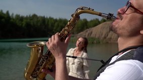 a young man in glasses and a black hat plays a saxophone near a forest lake. side view. in the background, a woman with long hair and a white dress is seen out of focus - Powered by Shutterstock - Get 15% off with code: PIKWIZARD15