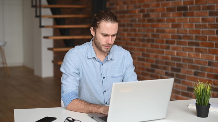 A male entrepreneur feeling tiredness, as a result of excessive exertion, lack of sleep, overworked with online project. A guy with laptop is massaging back of the neck, feels hurt and muscle tension | Shutterstock HD Video #1066365535