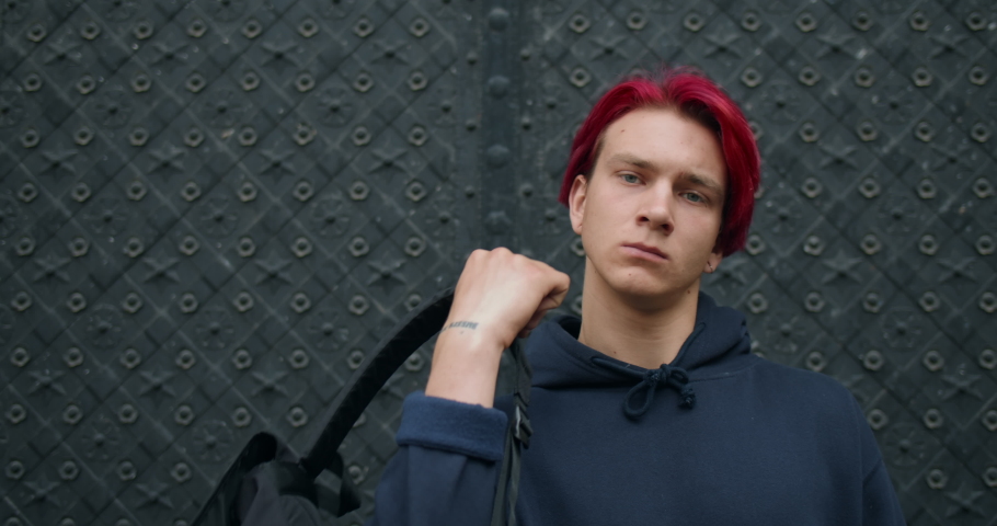 Portrait of millennial male hipster putting backpack on his shoulder while standing at street. Young guy with earrings and dyed red hair looking to camera. Concept of lifestyle