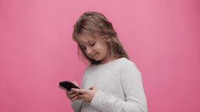 Portrait of happy little cute girl holding smartphone standing on pink background. Choosing favorite music or cartoons,texting messages,browsing internet,watching video, playing games on smartphone.