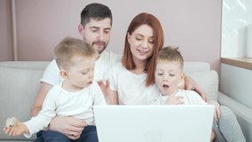 young happy family dad and mom with young children communicates online on laptop remotely. Waving hands and laughing sitting on the couch in the living room or kitchen at home talking to grandparents