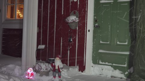 Two decorative snowmen - one plastic with light and another wooden with candle holder partly covered by fresh snow and standing close to green entry door of Swedish wooden red house. Show storm.