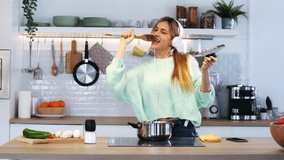 Video of beautiful young woman cooking while singing and listening music with headphones in the kitchen at home.